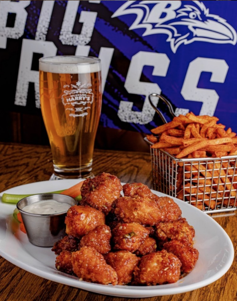 Image shows a plate of hot wings, a basket of fries, and a cold beer staged in front of a Baltimore Ravens sign. Mustang Harry's is one of the best bars in NYC to watch the Baltimore Ravens