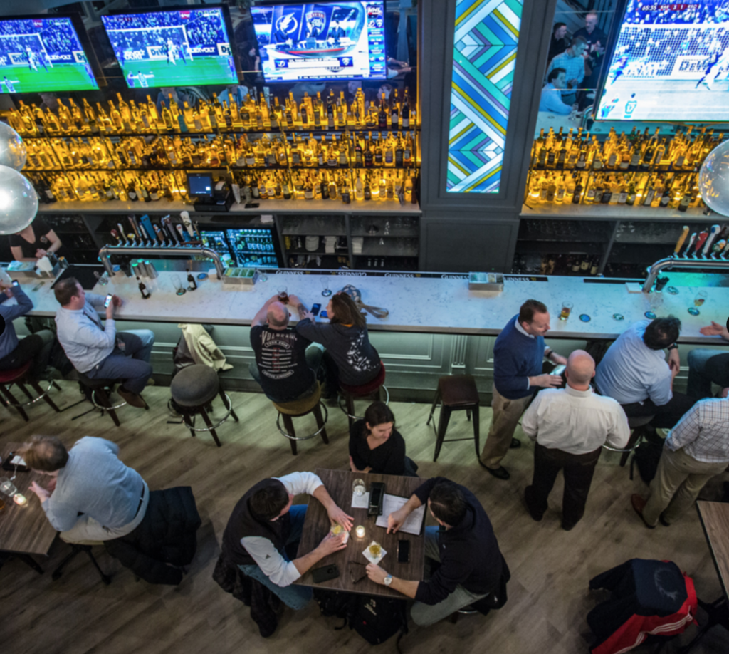 Image shows Tir Na Nog's illuminated bar accompanied by several monitors overhead displaying sports games. 